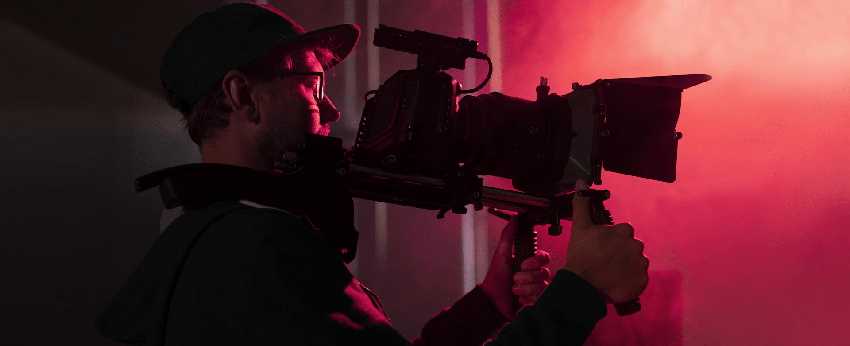 Operatore video riprende una scena con una videocamera professionale, illuminato da luci rosse e rosa.