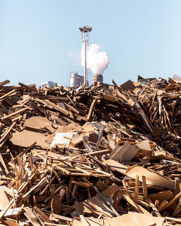 Cumulo di scarti di legno e cartone in un impianto industriale, con camino sullo sfondo.
