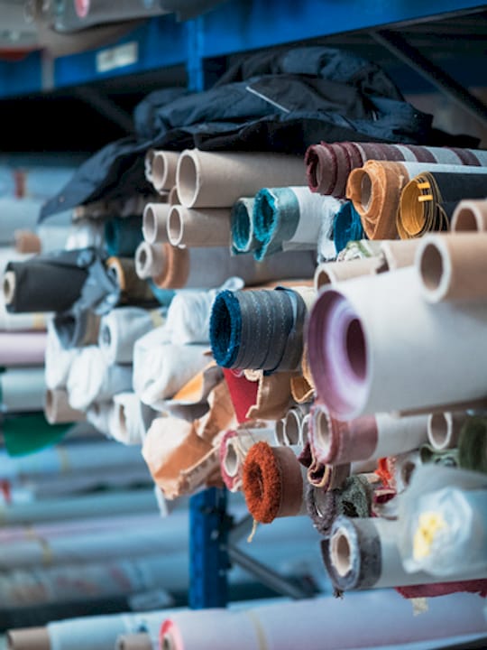 Rotoli di tessuti e materiali colorati disposti su scaffalature in un magazzino.